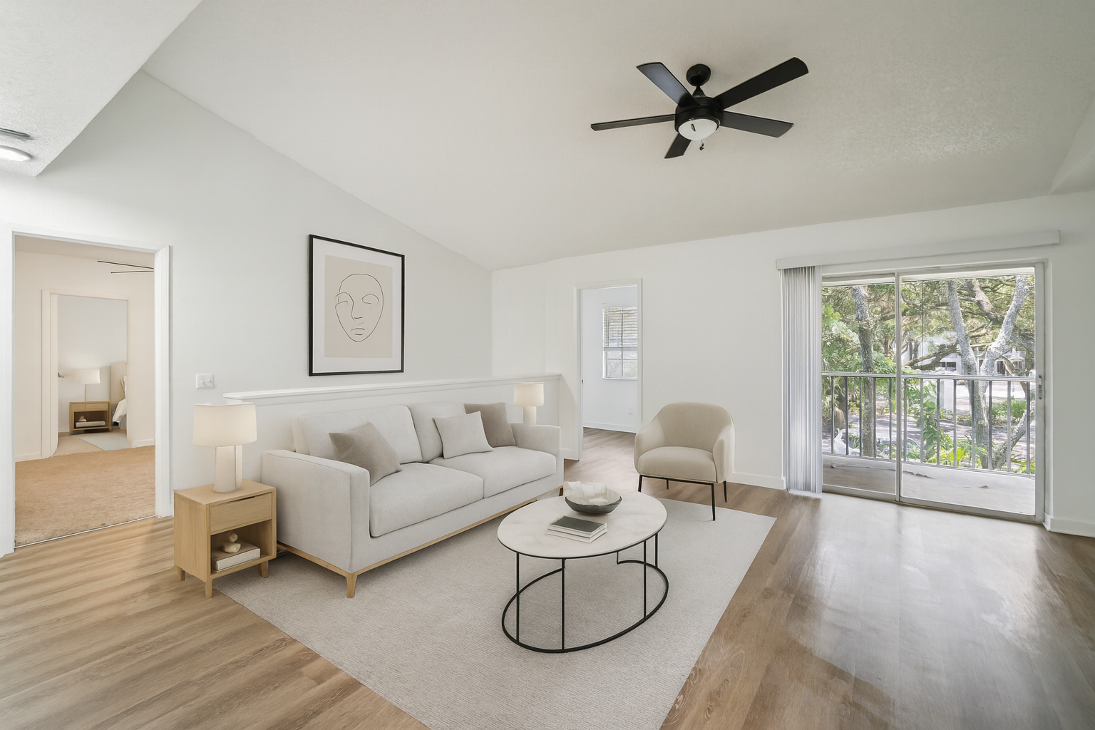 A serene and modern living room features a plush, light-colored sofa adorned with throw pillows, positioned centrally on a large area rug. The room is accented with a contemporary coffee table holding a few decorative items, and a stylish side table with a lamp. A comfortable armchair sits nearby, and a large framed artwork hangs above the sofa. The space is brightened by natural light streaming through a sliding glass door leading to a balcony, creating a tranquil atmosphere perfect for relaxation. The room's minimalist design and neutral color palette enhance its inviting and cozy ambiance.